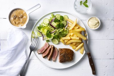 Een echte Vlaamse klassieker die smaakt voor jong & oud, een steak mooi gebakken met heerlijke goude frietjes en een salade. De champignonsaus staat langs het bord in een sauspotje. Bestek en servette liggen langs het bord klaar om te starten. 
