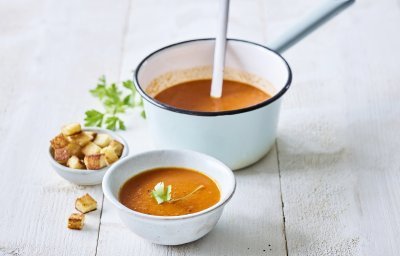 Op een houten witte tafel staat een kopje paprikasoep. De soepkom staat ook in beeld, dit is een lichtblauwe pot die mooi contrast geeft met de oranje-rode soep. De soep is afgewerkt met verse peterselie en croutons.
