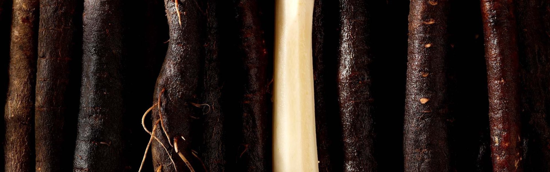 Sfeerbeeld van verschillende ongeschilde schorseneren. Hun bruine, bijna zwarte, kleur schittert onder de belichting. In het midden ligt 1 geschilde schorseneer, spierwit te blinken.