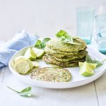 De pannenkoekjes van aardappel en spinazie liggen op een wit bord gestapeld. Er liggen blaadjes verse spinazie langs en partjes limoen. Op de witte houten tafel staan ook twee licht gekleurde blauwe glazen met water gevuld, een waterkan en een blauw linnen servette. 