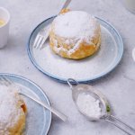 Heerlijke appelbollen staan klaar op tafel om toe te happen. Er staan twee kopjes kofie vij en verse jonagold appelen. De appelbollen zijn bestrooid met bloemsuiker. 