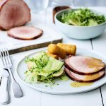Op een houten witte tafel staat een portie klaar. Een wit bord gevuld met twee sneetjes van het varkensgebraad, kroketten en de salade van komkommer met appel.