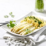 Op een rechthoekig wit bord liggen gekookte asperges, afgewerkt met geplette gekookte eieren, peterselie en gesmolten boter. Klaar om aan te vallen. 