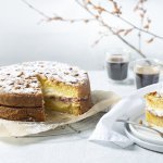 Een biscuit met vanillepudding en slagroom staat op tafel op een taartrooster. Er is al een stukje aangesneden om de lijnen duidelijk te laten zien van de pudding en slagroom. Koffie uitgeschonken, vorkje prikken en aanvallen die taart. 