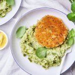 Op tafel staan 2 witte borden met een blauw randje. Hierin ligt een schep broccolipuree met een kaasburger en enkele blaadjes basilicum. Er staat ook een potje mosterd op tafel. 