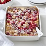 Op een witte houten tafel staat een rechthoekige ovenschotel met hierin de broodpudding klaargemaakt. Hij oogt zeer mooi door de toevoeging van het rode fruit en de afwerking met bloemsuiker. Er staan bordjes langs de ovenschotel om onmiddelijk op te scheppen en eventueel nog wat extra rood fruit te nemen. 