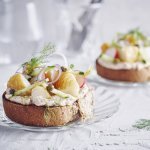 Een goedgevulde bruschetta ligt op een klein glazen bordje en staat op tafel. De bruschetta werd afgewerkt met een takje dille. Achteraan de tafel staat een kan met water en bestek om deze toast te verorberen. 