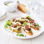 Op een houten, witte tafel staat een groot wit rond bord met hierop de bruschetta geserveerd. Ze zijn afgewerkt met extra blaadjes basilicum. Er staan ook kleine bordjes naast en de vijzel waarin de pesto is in gemaakt.
