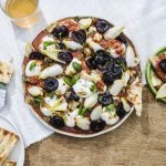Op een houte tuintafel staat een grote ronde witte serveerschaal. Hij is gevuld met de bolletjes burrata, gebakken witlof en rode vinagrette op basis van tomaten. Wij serveren hem met krokant naanbrood en gebakken kersen. 