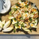 Op een houte snijplank staat een grote serveerschaal met hierin de caesarsalade geserveerd. Er staan enkele bordjes langs met bestek om onmiddelijk te starten. 