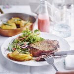 Een hoofdgerecht om U tegen te zeggen. We serveren een malse chateaubriand met millefeuille van aardappelschijfjes en knapperige kruidensalade. De frambozendressing staat langs het bord in een klein glazen flesje. Er staat ook een glas water boven het bord, naast peper en zout.