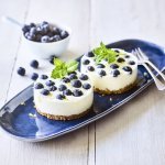 Op een witte houte tafel staat een langwerpig blauw bordje. Hierop liggen twee mini-cheesecakes met blauwe bessen. Er ligt een vork langs, een potje extra blauwe bessn en een takje munt.