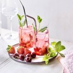 Twee cocktails staan op tafel, ze hebben een mooie roodroze kleur. Onderaan het glas ligt het smached fruit, met daarna de vodka en roze tonic. Er liggen nog enkele aardbeien en kersen langs. 