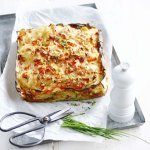In een ovenschotel ligt een geweven taartje van courgette en aardappel met stukjes rode paprika. Hij heeft mooie kleuren van geel, rood en groen door de verse bieslook. 