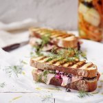 Twee croques liggen op het aanrecht in de keuken op een wit boterpapiertje. In de achtergrond staan de gepekelde groentjes (wortel en komkommer) in een bokaal. De tosti's zijn mooi gegrild in streepjes.