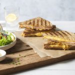 Een krokant gebakken croque monsieur ligt aangesneden op een houten snijplank. Lang staat een salade in een diep wit bord met een potje mayonaise voor te dippen. 