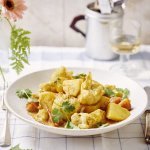 Heerlijke goudkleurige curry geserveerd in een diep wit bord met bloemkool en aardappelen. Er ligt bestek langsheen het bord en glaasje witte wijn. Ook zien we een vaasje met roze bloem en varentakje. 