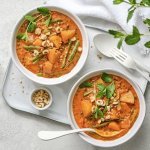 Twee heerlijke curry staan uitgeschept op tafel. Ze hebben een mooie oranje kleur door, jawel, de curry pasta. Ze staan beiden op een poseleinen dienplateau met bestek en een potje pina- en hazelnoten. 