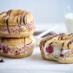 Drie donuts liggen op een witte stenen snijplank. De donuts zijn doormidden gesneden en gevuld met yoghurtijs met rode framboosjes, gerold door kleurrijke sprinkles en afgewerkt met gesmolten chocolade. In de achtergrond staat nog een glaasje melk.