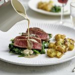 Een wit bord staat op tafel met hierop de heerlijke krokante aardappel en de saignant gebakken filet pur op een bedje van spinazie. Er komt een sauskannetje in beeld die saus over het vlees giet. De tafel is gedekt voor twee met de perfecte maaltijd.