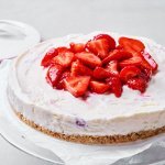Een heerlijke cheesecake staat op een glazen serveerschaal. Dit is niet zomaar een cheesecake, maar eentje die diepgevroren is. Perfect voor een koel dessertje op een zomerse dag. Er liggen enkele verse aardbeien op de taart om het fruit extra in de verf te zetten, want aardbeien zijn gewoonweg de toppers van het zomerfruit. 