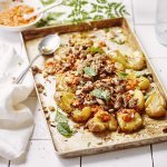 Op een ovenschaal liggen krokante crushed potatoes met homemade romesco saus bovenop en afgewerkt met lamsgehakt. De overige romesco saus staat bovenaan voor wie wat extra wil. Er ligt een witte servette op het aanrecht van witte Marokaanse tegeltjes. 