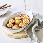 Witte ovenschotel staat op een houten snijplankje op tafel. De ovenschotel is gevuld met de rode tomatensaus waarin de champignons mooi in geschikt zijn. Achteraan de foto zie je bordjes staan met bestek om aan de slag te gaan. 