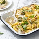 Op tafel staat een ovenschotel met hierin de pasta, prei, broccoli en kaas. Hij is gegratineerd in de oven, wat een mooi goud korstje geeft en afgewerkt met verse peterselie. Er is al een portie uitgeschept en het bordje staat boven de shotel. 