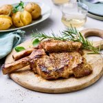 Gegrilde koteletten liggen op een houten ronde snijplank te blinken. Er ligt een takje rozemarijn langs, een bordje met de gegrilde krieltjes en een potje met de salsa verde om toevoegen aan het vlees.