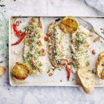 3 stukjes gegrilde vis liggen op een rechthoekige ovenschaal. De gegrilde citroen en chilipepers zorgen voor kleur in dit gerechtje, samen met de groene kruiden in de crumble en dip. 