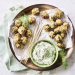 Op het aanrecht staat een houten ronde serveerschaal met een wit boterpapiertje op. Hierop liggen kleine gehaktballetjes met groene stukjes in van de courgette en spinazie. De dip op basis van tzatziki staat ook op de schaal. Het gerecht is afgewerkt met enkele takjes dille. 