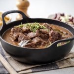 Er staat een zwarte sudderpot op tafel met Gentse stoverij, in de achtergrond zijn er frietjes te zien en een witloofslaatje.