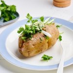 Een gepofte aardappel ligt op een bordje, gevuld met homemade kruidenboter, lente-ui en verse kruiden. 