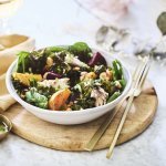 Een salade van bietjes, boerenkool en spinazie afgewerkt met noten en makreel. Deze feestsalade staat op een houten plank met de dressing in een apart gouden potje.