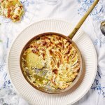 Een heerlijke gratin met groenten en kaas klaargemaakt in een ovenbestendige pan staat in het midden van de tafel. Er is al een portie uitgeschept op een bordje. Op tafel ligt een wit tafelkleed met enkele geborduurde blauwe bloemen. 