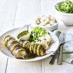 Op een bleke houten tafel staat een diep bord met hierin een schep aardappelsalade in, twee groentespiesjes met schijfjes van gegrilde aubergine, courgette, champignons en een kannetje met rucolapesto. Verder staat er ook nog een kom met verse groene salade, bestek en een muntkleurige servette.