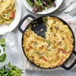 Een taart van aardappelpuree en paprika, staat op het midden van de tafel. Hij zit in een zwarte schotel en heeft een mooi kaaskorstje meegekregen uit de oven. Er is al 1 portie uitgeschept, en er ligt ook bestek op tafel, sla en verse kruiden. 