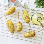 Hasselback potatoes liggen op een bakrooster te blinken. Ze zijn doorprikt met lange houten prikker om makkelijker te kunnen draaien op de BBQ. Er staat een flesje olie, verse tijm en een potje zeezout naast. 