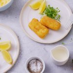 Twee witte bordjes staan op een licht grijze tafel, op elk bordje liggen twee kaaskroketten met partjes citroen en verse tuinkers. 