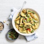Er staat een grote witte serveerschaal op tafel gevuld met de goudgele kip curry. Je ziet de groene peultjes en rode chili duidelijk. Langs het gerecht staan nog extra noten, gesneden lente-uitjes en een bordje rijst. 