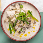 Kip gegaard in melk staat op tafel in een diep beige bord met bordeau randje. Er liggen lente-uitjes en champignons bij. Het bordje staat op een blauwgroene onderlegger. 