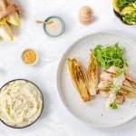 Gestoofd witloof ligt op een wit bord, samen met de kipfilet en mosterdsaus. De groene rucola zorgt voor een frisse kleur in dit gerecht. 
