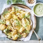 Puree van knolselder is uitgeschept op een groot lichtbruin bord. Hierop zijn de geroosterde bloemkoolroosjes geserveerd met wat drizzle van de groene kruidendressing, noten en verse dille. De overige kruidendressing is in een witte potje en staat langs het bruine bord, samen met nog wat gehakte noten in een ander potje. Alles staat op een een plaid in een blauwgrijze kleur.