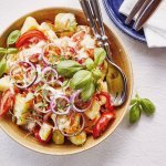 Een grote serveerschaal staat op de tafel, ze is gevuld met gekookte aardappelstukjes, tomaat, paparika, rode ui en wortel. De dressing is al toegevoegd en verse kruiden zoals basilicum en tijm geven een nog extra kleurtje aan de salade.