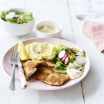 Een homemade shnitzel ligt op een pastelroze bord, samen met aardappelpuree, verse sla en zelfgemaakte mayonaise. Er is nog wat extra salade in een wit potje en mayonaise voor extra lekkers op te scheppen. 