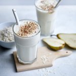 Twee klaargemaakte milkshakes staan op het lichtgrijze aanrecht te pronken. Er ligt een peer langs die doormidden gesneden is en wat havermoutvlokken gemorst. De shakes zijn afgewerkt met wat kaneelpoeder. 