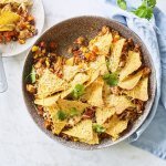 Onderaan de ronde ovenschaal ligt de chili con carne met bovenop de nachos. Er is al een portie uitgeschept op een wit bordje en staat langs de ovenschaal. Er ligt ook een blauwe servette en enkele blaadjes kroiander voor de kleur.