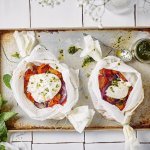 Twee papillotten liggen op een ovenplaat. De papillot is gemaakt in een wit boterpapiertje waarin de groenten een mooi contrast in geven. Denk maar aan de rode paprika's en tomaten. De burrata ligt er bovenop en de muntolie staat in een bokaaltje naast de papillotten.