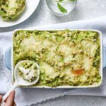 Eten is klaar! De ovenschotel staat op een witte dikke doek en een porseleine lepel schept de eerste portie uit de schotel. De parmentier heeft een mooie frisse groene kleur door de vele erwtjes. 