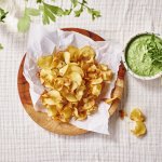 Op tafel ligt een kalkwit tafelkleed, met hierop een houten bordje met wit boterpapiertje en chips van pastinaak op. Er staat een dippotje langs met de dip van broccoli en roomkaas. 
